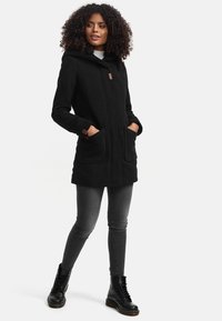 Black hooded coat with two large front pockets, featuring a smooth texture and contrasting brown accents. Paired with grey jeans and black boots.
