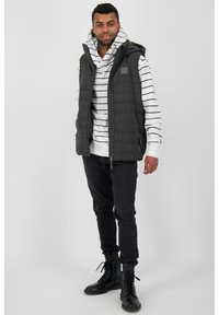 Gewatteerde, donkergrijze vest met capuchon, gedragen over een wit en zwart gestreept sweatshirt. Zwarte jeans en combatboots maken de outfit compleet.
