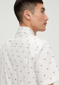 Herren Kurzarmhemd aus weißem Stoff mit einem kleinen, sich wiederholenden Muster aus roten und grauen Akzenten. Klassischer Kragen und glatte Textur.