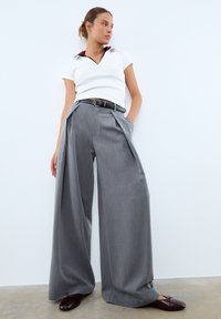 Femme portant un pantalon gris large, un haut blanc à manches courtes, une ceinture noire et des ballerines rouge foncé, se tenant contre un mur blanc.