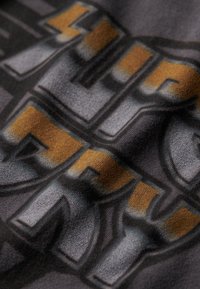 Tessuto grigio con una superficie testurizzata caratterizzato da grandi lettere in oro e nero. Il design include ombreggiature per profondità.