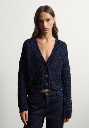 Joven mujer vestida con una chaqueta de punto grueso azul marino con botones sobre pantalones oscuros de cintura alta, de pie contra un fondo liso.