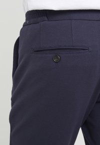 Mørkeblå bukser med tekstureret stof, flad front, elastisk talje og en enkelt baglomme med en sort knaplukning.