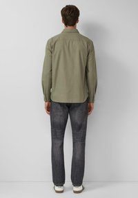 Man die naar een muur kijkt en een olijfgroene langslavende shirt, donkergrijze jeans en witte sneakers met zichtbare merkl logo draagt.