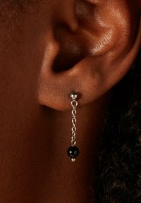 Les boucles d'oreilles en chaîne argentée présentent un pendentif en perle noire, mettant en avant un design simple avec une finition polie, suspendu élégamment au lobe de l'oreille.