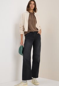 Lichtbeige bomberjack over een bruine hoodie, gecombineerd met zwarte wijde jeans en beige sneakers. Houdt een groene pet vast en staat tegen een neutrale achtergrond.