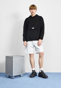 Felpa nera con cappuccio e tasca frontale, pantaloni corti grigi e sneaker nere. Un'unità di stoccaggio mobile in metallo argentato accanto a un tappeto blu.