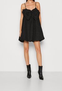 Svart klänning med en stor rosett fram, gjord av strukturerat material, med tunna remmar och en utsvängd silhuett. Buren med ankelstövlar.