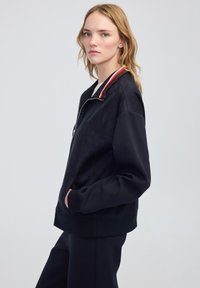 Chaqueta bomber azul marino con cierre frontal, que presenta un cuello de canalé con acentos en rojo y blanco, tejido texturizado y bolsillos laterales.