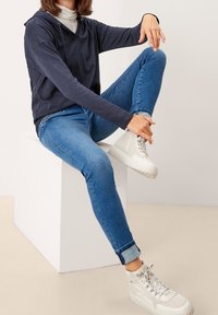 Tmavě modrá mikina s kapucí a dlouhými rukávy, přiléhavé modré džíny a bílé tenisky sedící na bílém podstavci na neutrálním pozadí.