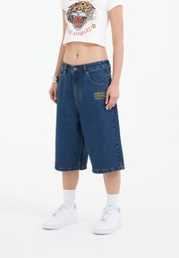 Denim shorts in dark blue, knee-length, with yellow embroidered logo detailing. Worn with a cropped white graphic t-shirt and white sneakers.