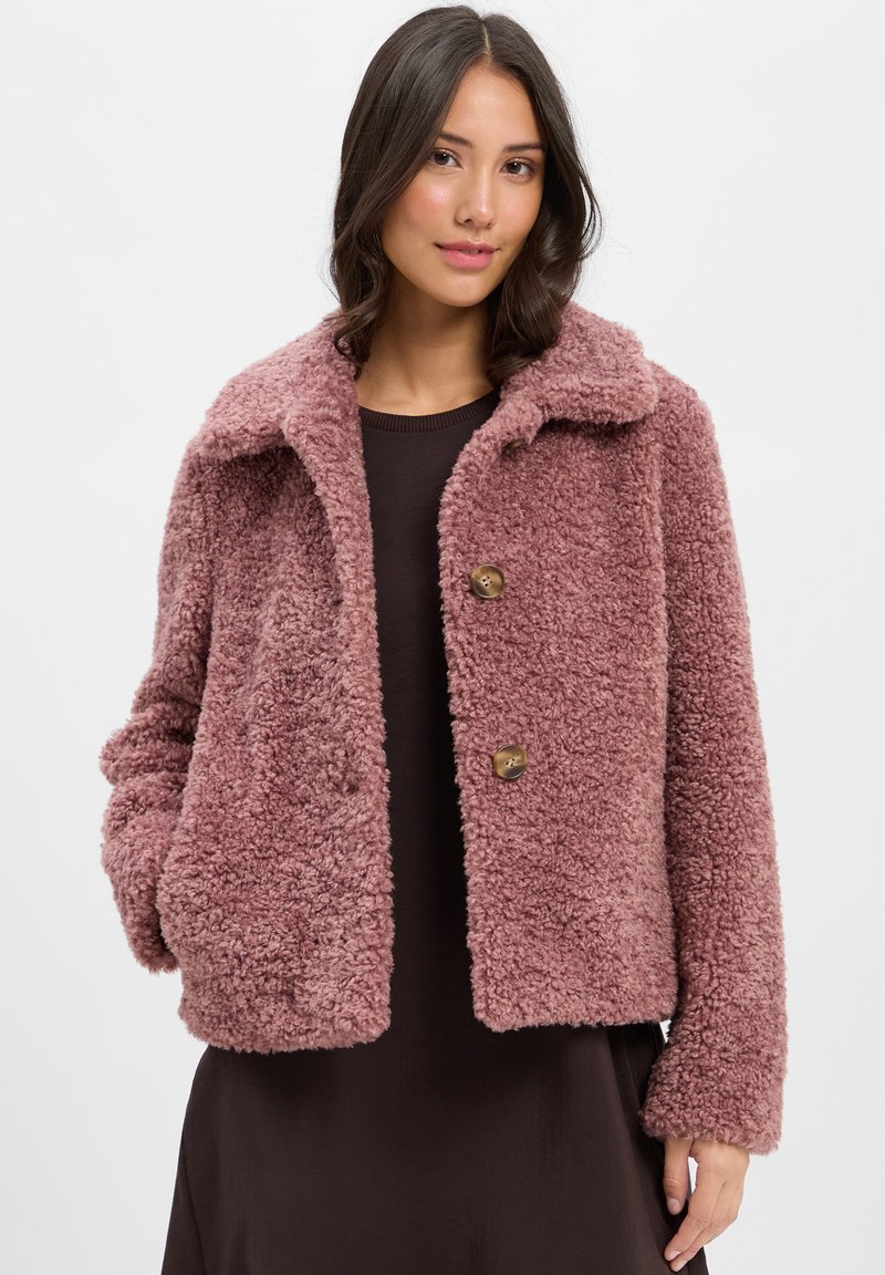 Frau in einem mauvefarbenen, strukturierten Teddy-Mantel mit Knöpfen über einem dunklen Kleid, stehend vor einem schlichten, hellen Hintergrund.