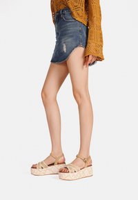 Sandali con zeppa e cinturini intrecciati in beige, con una base platform a fantasia, indossati con una gonna di jeans e un maglione di maglia color senape.