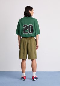 Green mesh jersey with black number "20" on back, paired with olive green shorts. Model wears red, white, and black sneakers with white socks.