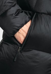 Schwarze Pufferjacke aus Stoff mit glatter, glänzender Textur; verfügt über eine Reißverschlusstasche, diagonale Nähte und ein Rautenmuster.
