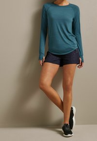 Tastgrün langärmliges Sportoberteil mit strukturiertem Muster, kombiniert mit dunkelblauen Shorts. Das Model trägt schwarze Sneakers mit weißen Sohlen.