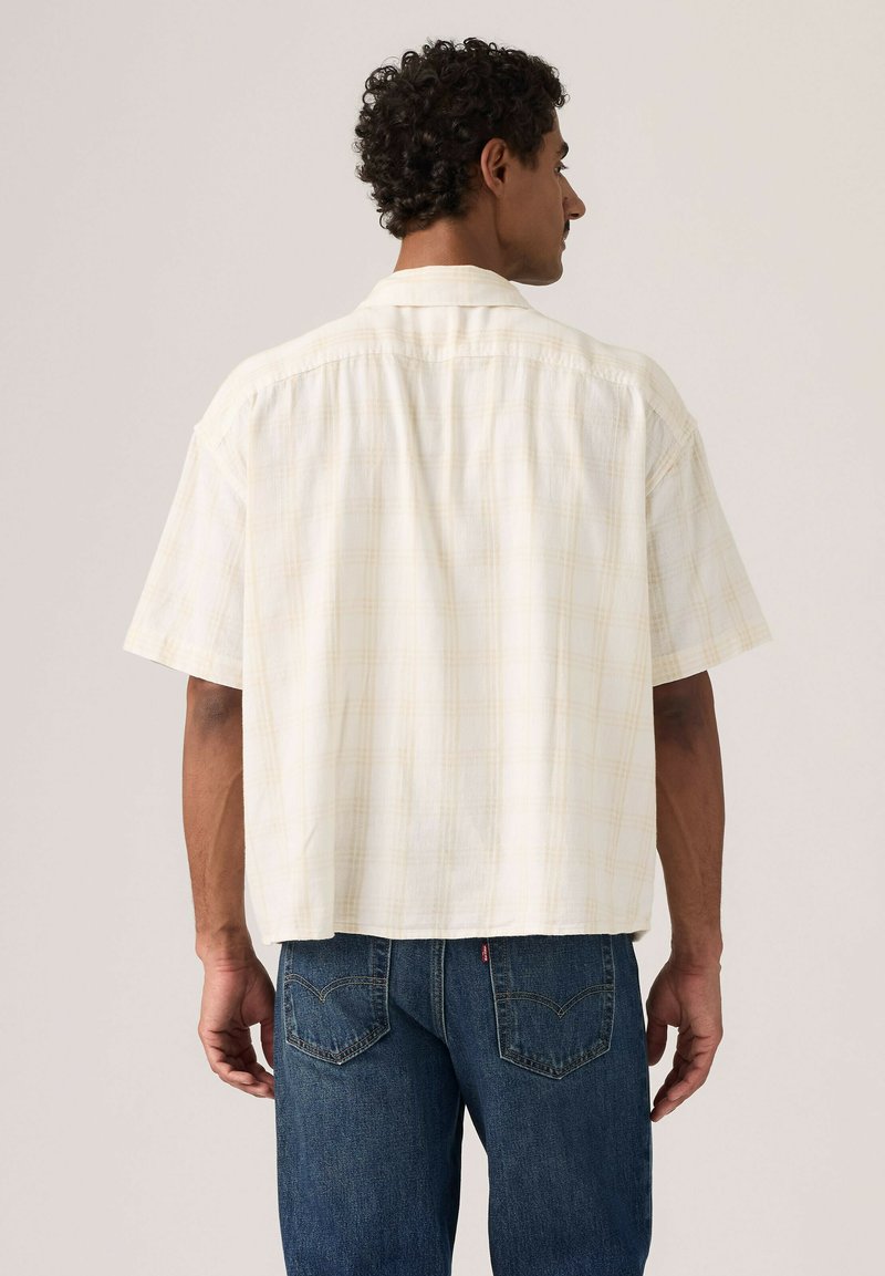 Mann trägt ein locker sitzendes, kurzärmeliges, cremefarbenes kariertes Hemd und blaue Jeans, von hinten vor einfarbigem Hintergrund gezeigt.