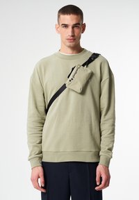 Hellgrüner Pullover mit langen Ärmeln, Rundhalsausschnitt und einer schwarzen Umhängetasche mit Logo. Die Tasche ist rechteckig und hat eine strukturierte Oberfläche.