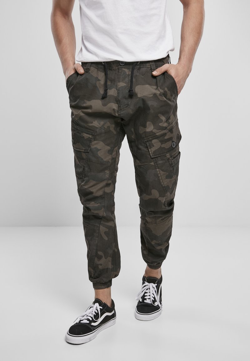 Homme portant un jogging cargo camouflage foncé et des baskets noires à lacets blancs, debout les mains dans les poches devant un fond uni.