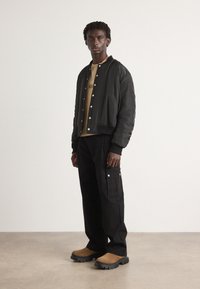 Black bomber jacket with silver snap buttons over a tan shirt, paired with black cargo pants featuring pockets and brown, rugged slip-on shoes.