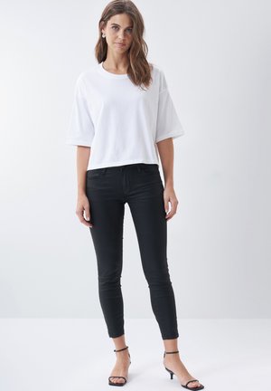 Bílý bavlněný tričko s krátkým rukávem, zkrácený design, nošené s černými skinny džíny a černými sandály na podpatku, na neutrálním pozadí.