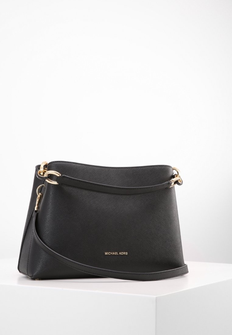 Sac à main noir Michael Kors avec des éléments dorés et deux anses, posé sur une surface blanche sur un fond uni et clair.