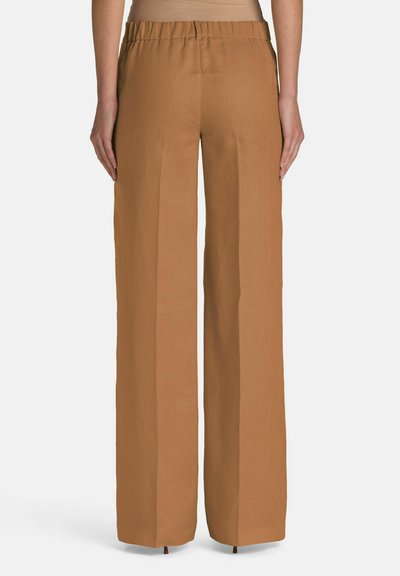 Vrouw die een camel-kleurige broek draagt met hoge taille en brede pijpen, met een elastische tailleband, staand met de armen ontspannen langs het lichaam, gezien van achteren.