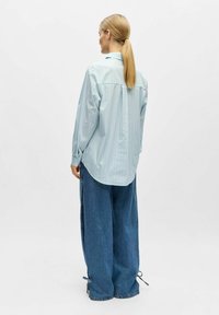 Chemise à rayures bleu clair avec boutons, manches longues, et jean large et ample avec liens aux poignets. Vue de dos.