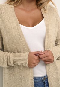 Femme portant un cardigan en tricot beige sur un haut blanc à col en V et un jean bleu, les mains jointes au niveau de la taille.