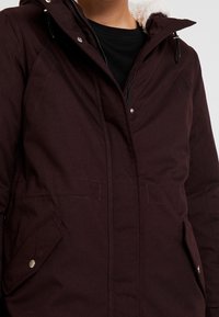 Giacca parka bordeaux con chiusura a zip, tasche con bottoni a pressione e fodera del cappuccio morbida e lavorata. Presenta un tessuto resistente e cuciture visibili.