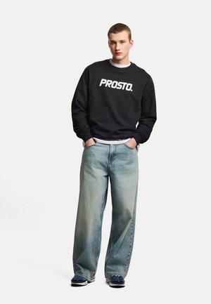 Młody mężczyzna stoi z rękami w kieszeniach, ubrany w czarną bluzę "PROSTO.", jasnoniebieskie szerokie spodnie dżinsowe i niebieskie trampki.