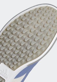 Gros plan de la semelle d’une chaussure Adidas avec des motifs triangulaires texturés et en relief pour l’adhérence, et un tissu bleu visible au bord.