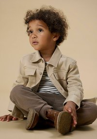 Malé dítě s kudrnatými vlasy sedící s překříženýma nohama, oblečené v béžové bundě, pruhovaném tričku, hnědých kalhotách a hnědých botách, které se dívá lehce vzhůru.