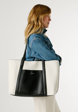 Mujer con chaqueta vaquera que lleva un gran bolso tote blanco y negro con asas largas sobre el hombro, mirando hacia la derecha.