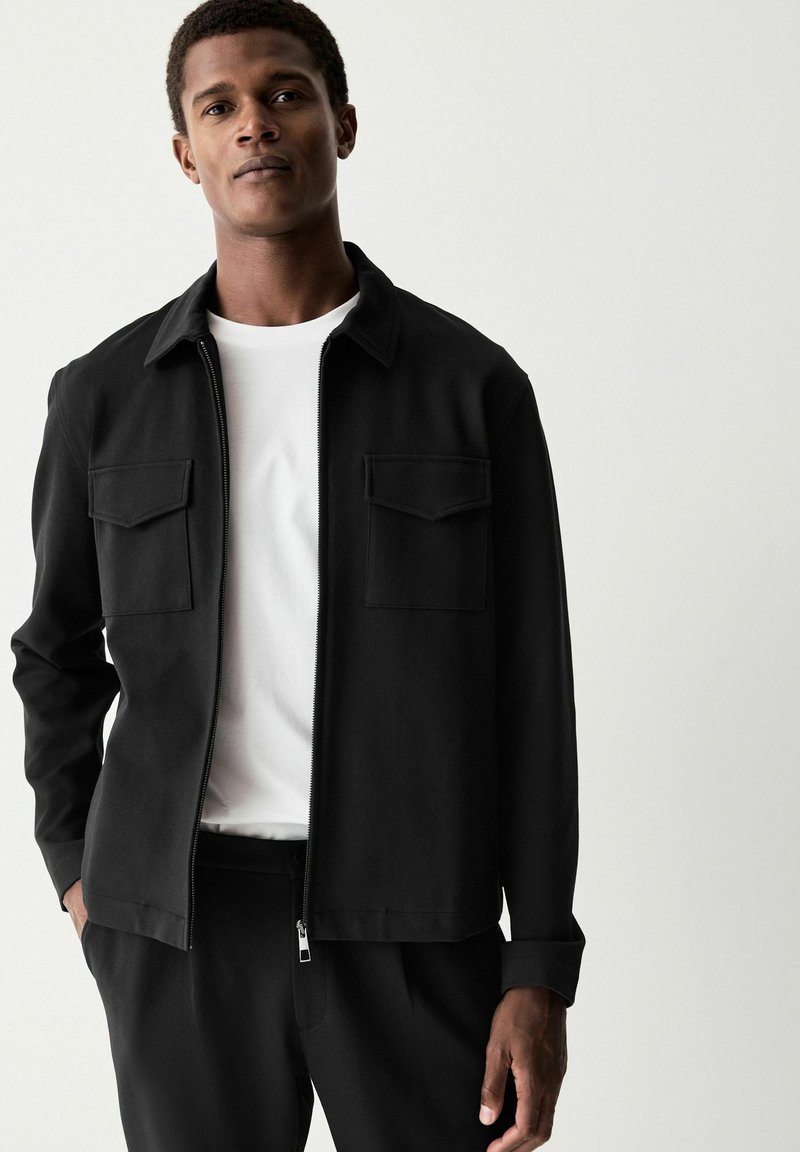 Chaqueta negra fabricada en tejido suave, con cierre de cremallera en el frente, dos bolsillos en el pecho y un collar. Combinada con una camiseta blanca y pantalones negros.