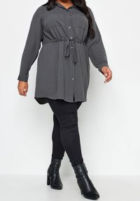 Grijze button-up overhemdjurk met een aantrekkoord in de taille, lange mouwen en zakken. Gepaard met zwarte skinny jeans en enkellaarsjes.