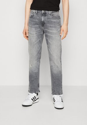 Personne portant un jean gris délavé droit et des baskets blanches avec des rayures noires sur un fond uni.