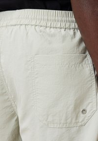Light beige shorts with an elastic waistband, single rear pocket with a rounded flap, and a silver grommet detail. Soft, textured fabric.