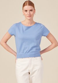 Top blu in cotone a maniche corte con scollatura rotonda e vita aderente, abbinato a pantaloni bianchi. Colore uniforme, texture liscia.