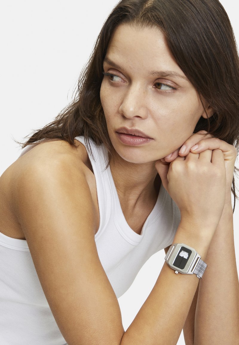 Silverfärgad metallarmbandsur med en rektangulär svart urtavla, nätarmband och minimalistisk design, bärs på en persons handled.