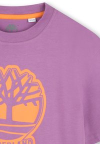 Maglietta di cotone viola con scollo rotondo e grafica di un logo ad albero arancione sul davanti. Maniche corte e texture liscia.