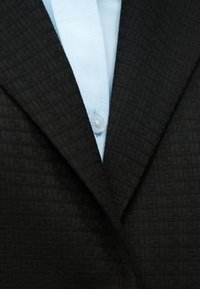 Blazer noir texturé porté sur une chemise bleu clair boutonnée, montrant le col et les détails des boutons.