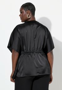 Top in satin nero con maniche corte a kimono, vita sottomessa e texture liscia, visto da dietro.