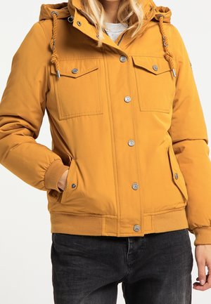 Donna che indossa una giacca con cappuccio color senape con bottoni a pressione sul davanti e tasche con patta, abbinata a jeans neri, in piedi con una mano in tasca.