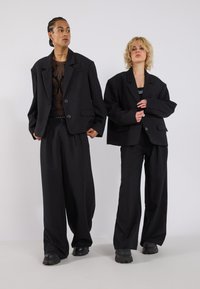 Twee modellen dragen oversized zwarte blazers met grote knopen, gecombineerd met wijde zwarte broeken. De stof lijkt glad en gestructureerd.