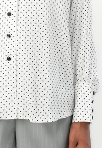 Weiße Bluse mit langen Ärmeln, schwarzen Punkten und schwarzen Knöpfen, die mit hellgrauen Nadelstreifenhosen von einer dunkelhäutigen Person getragen wird.