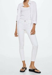 Jeans blancos y camiseta ajustada combinados con una camisa blanca de botones extragrande. La modelo lleva chanclas negras, posando frente a un fondo liso.