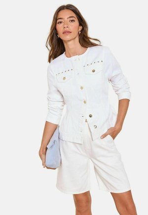 Kobieta ubrana w białą kurtkę zapinaną na guziki z ćwiekami oraz białe szorty, trzyma jasnoniebieską kopertówkę w jednej ręce, druga ręka w kieszeni.