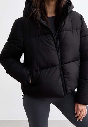 Mujer con chaqueta acolchada negra con capucha y leggings negros, de pie con una mano en el bolsillo y la otra apoyada al costado.