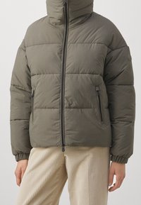 Person in olivgrüner Steppjacke mit Frontreißverschluss und mit Reißverschlüssen versehenen Taschen, kombiniert mit beigen Cordhosen.